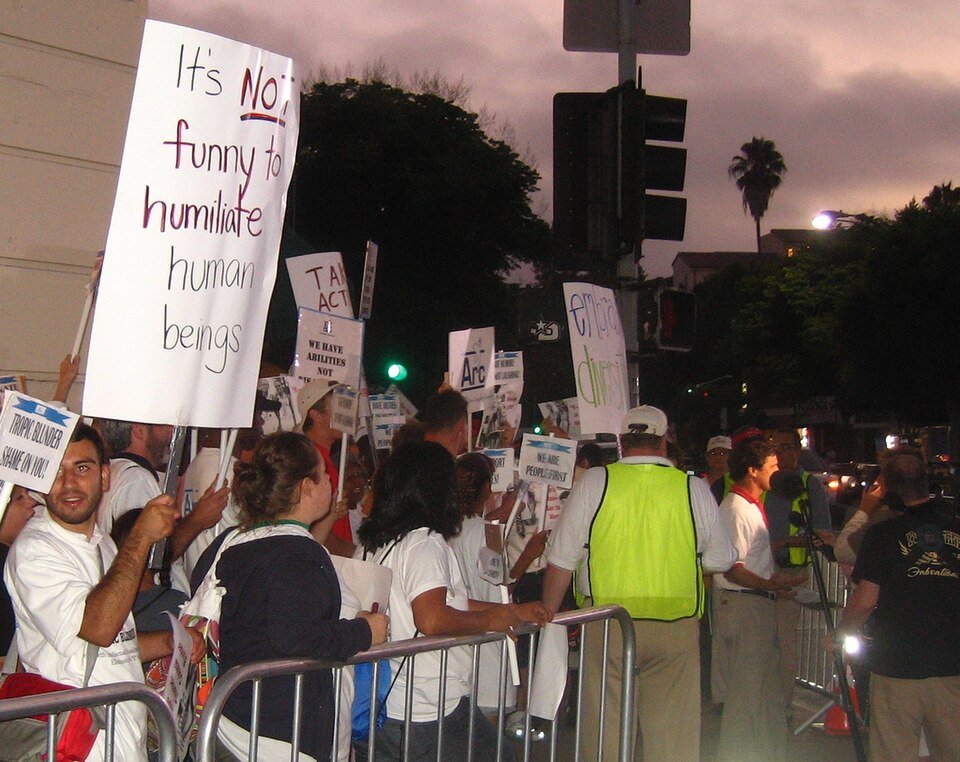 A group of protesters stand behind metal barricades at dusk, holding signs with messages such as Its NOT funny to humiliate human beings. Most people wear white shirts, and several officials in yellow vests are present.