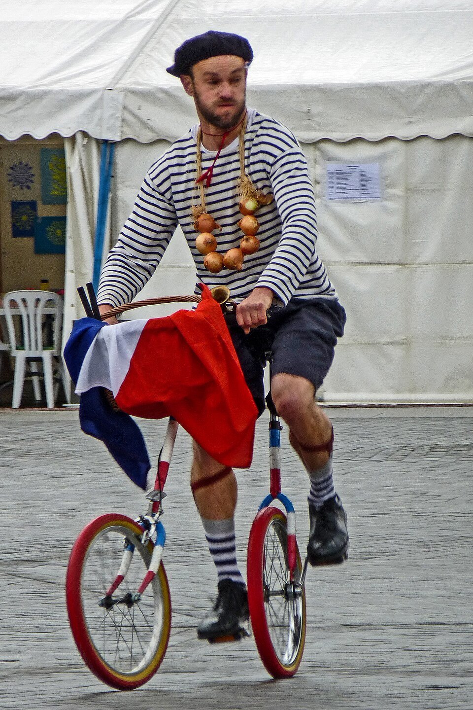A man in a striped shirt, beret, and onion necklace rides a bicycle with a French flag draped over the handlebars. He is in front of a white tent and appears to be performing or in a festive setting.