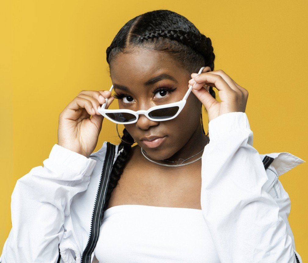 A young woman with braided hair wearing white sunglasses and a white outfit poses against a yellow background, looking over the top of her sunglasses with a confident expression.