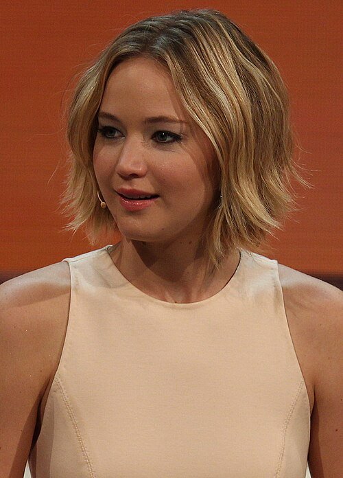A woman with short, wavy blonde hair is wearing a sleeveless, light beige top. She is looking slightly to her left against a warm, orange-brown background.