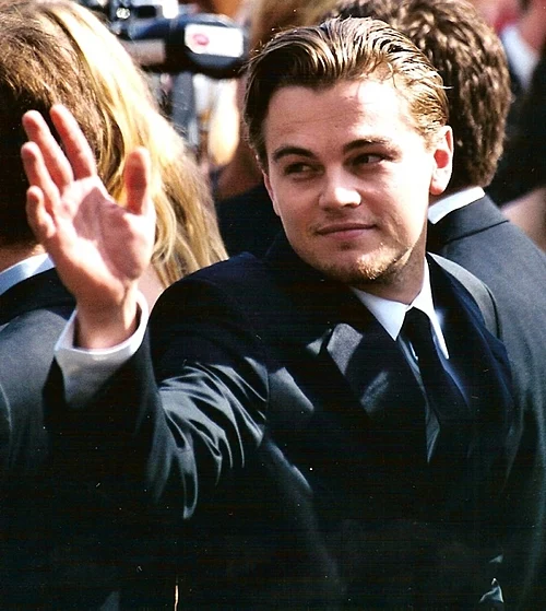 A man in a suit waves and smiles while standing among a crowd. His hair is neatly combed back, and the background is filled with people and cameras, suggesting a formal or public event.