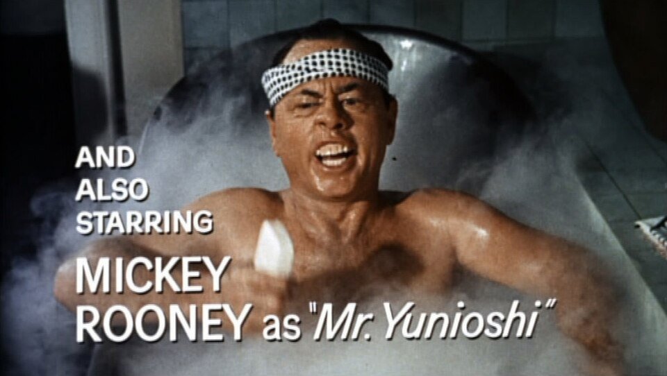 A man with a patterned headband sits in a steaming bathtub, grimacing and holding a brush. White text on the image reads: “AND ALSO STARRING MICKEY ROONEY as ‘Mr. Yunioshi’.”.