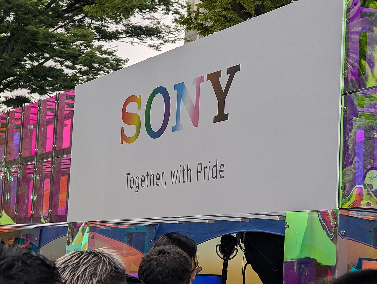 A large sign displays the Sony logo in rainbow colors above the slogan Together, with Pride. Iridescent decorations and a crowd of people are visible in front, with trees in the background.