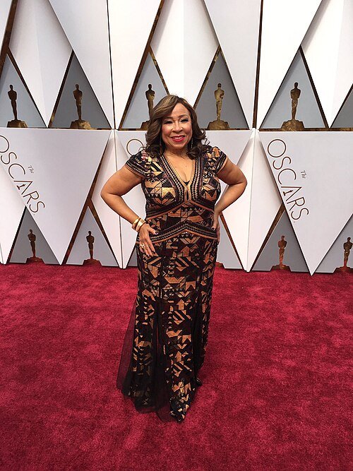 A woman in a black and gold embroidered gown stands on a red carpet at the Oscars, posing with one hand on her hip in front of a geometric backdrop featuring Oscar statuette designs.