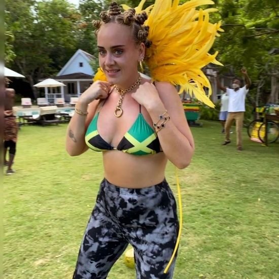 A woman wearing a bikini top with the Jamaican flag, tie-dye leggings, and yellow feathered wings stands on grass outdoors. She has bantu knots in her hair and is smiling. There are trees, houses, and people in the background.