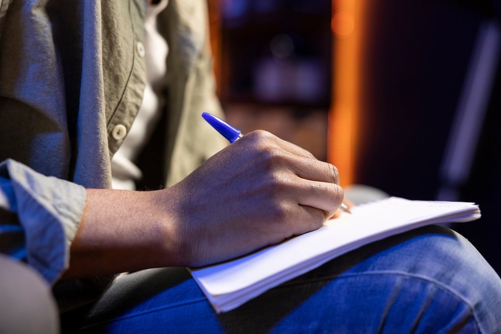 A person wearing a green shirt and blue jeans is sitting and writing in a notebook with a blue pen. The focus is on their hand and the notebook.