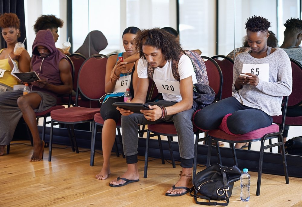 A group of people with numbered audition tags sit on chairs in a waiting area, reading or using their phones, appearing focused and relaxed.