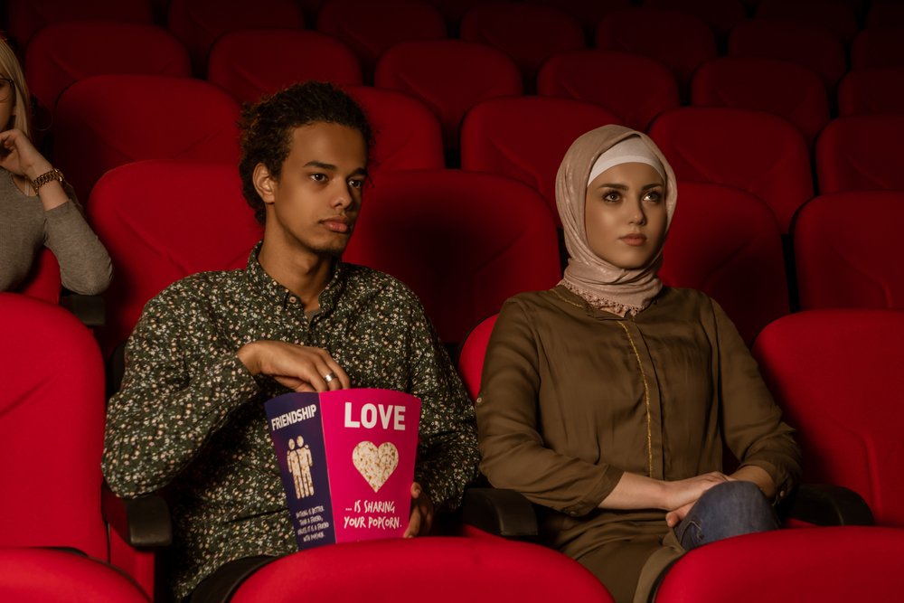 A man and a woman sit side by side in a movie theater with red seats. The man holds a popcorn box labeled LOVE and FRIENDSHIP. Both appear serious and are focused on the screen.