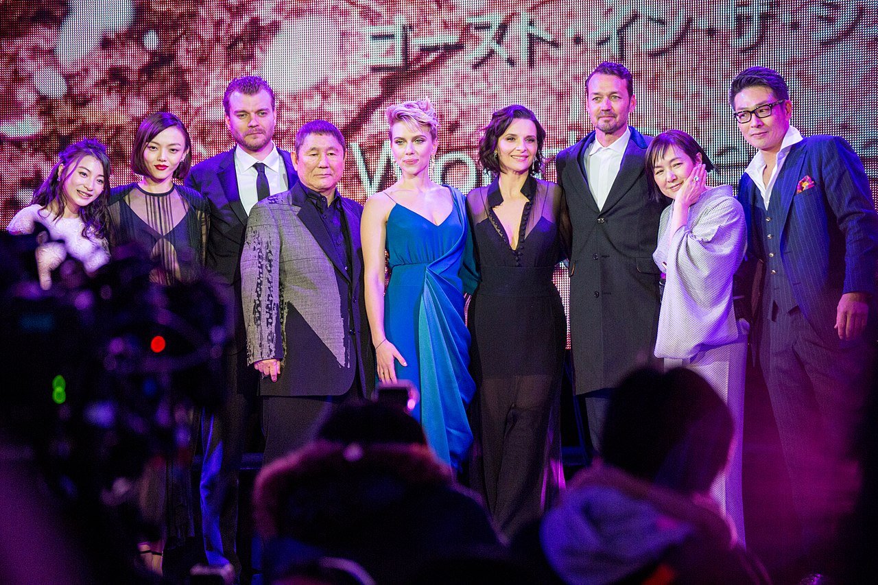 A group of nine people, dressed in formal and semi-formal attire, pose together on a stage with colorful lighting and a backdrop featuring Japanese text. An audience and camera equipment are visible in the foreground.