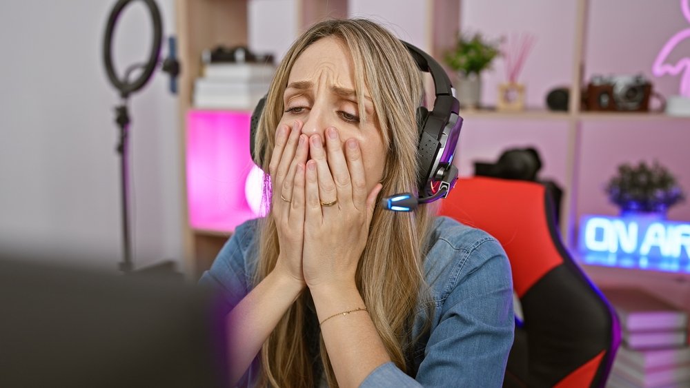 A woman wearing a headset sits in a gaming chair with her hands covering her mouth, looking distressed while staring at a computer screen. Shelves with decor and tech equipment are in the background.